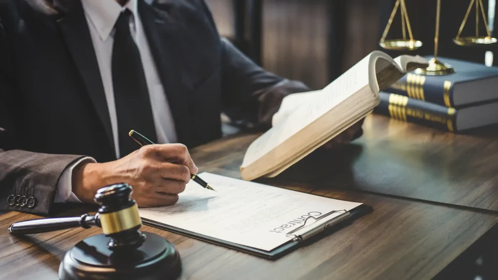 A lawyer signing documents while referring to a legal book, with a gavel in the foreground.