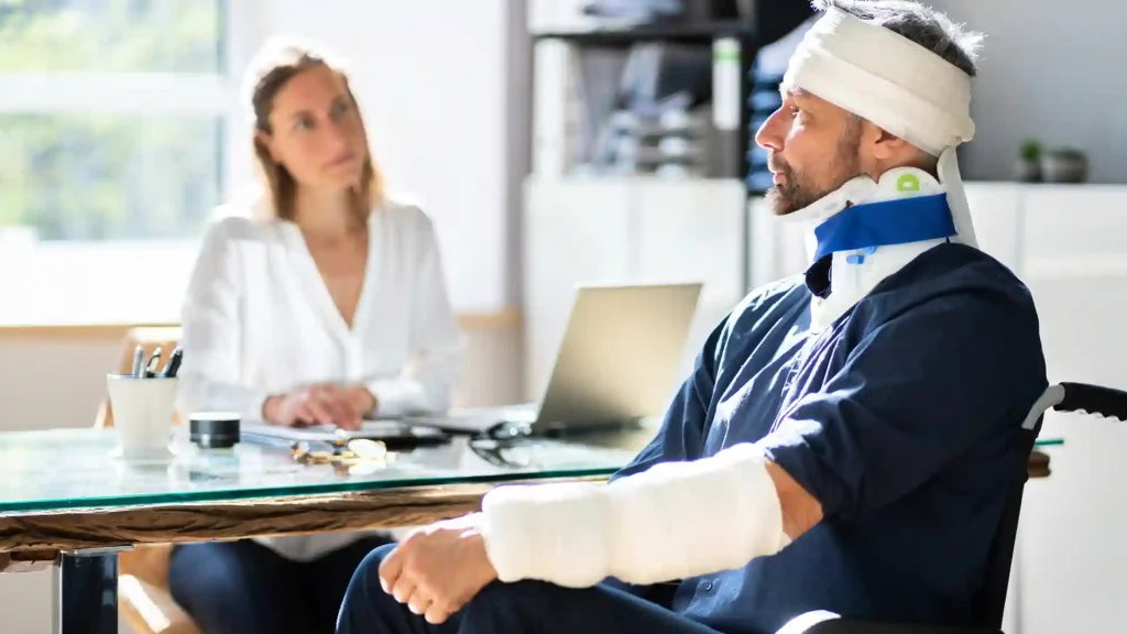 A man with bandages on his arms, neck, and head is sitting across from a female doctor during a consultation about his injuries