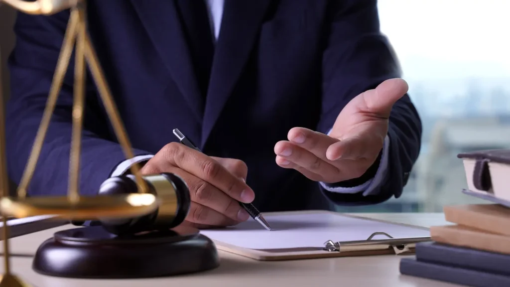 Attorney in a dark suit gesturing while taking notes during a legal consultation, with a gavel visible on the desk.