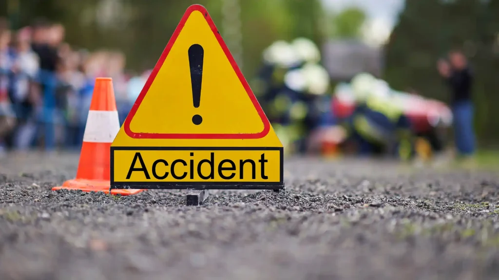 Yellow warning triangle sign with the word 'Accident' placed on a road surface, with emergency responders blurred in the background.